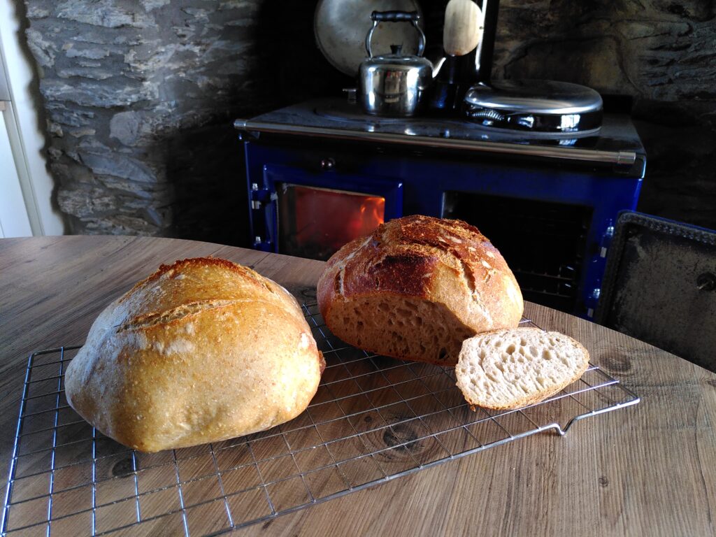 Freshly baked sourdough bread on a cooling rack. The bread is on the countertop. A wood-fired range cooker is on in the background.