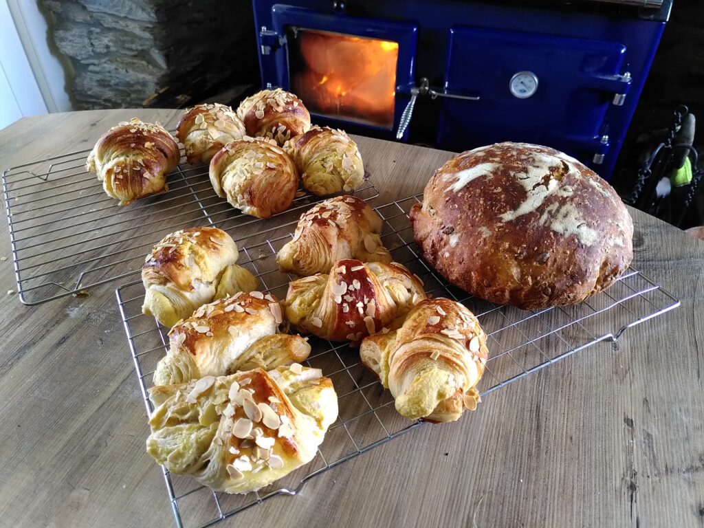 Freshly baked sourdough bread and homemade almond croissants, on a cooling rack in front of a wood-burning range in a farmhouse kitchen