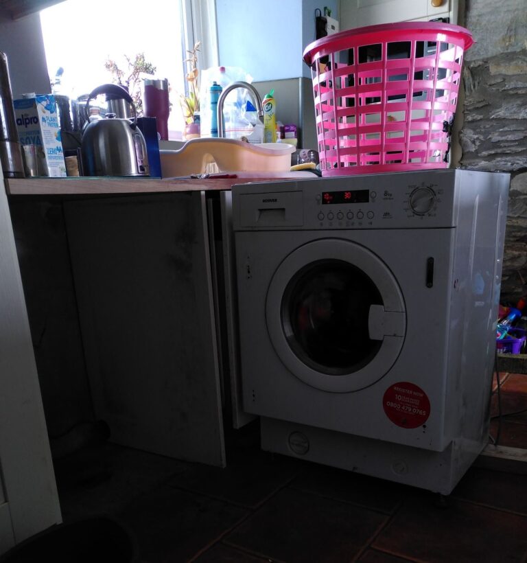 A farmhouse kitchen. The washing machine has been extracted from under the counter top and is in the middle of the room.