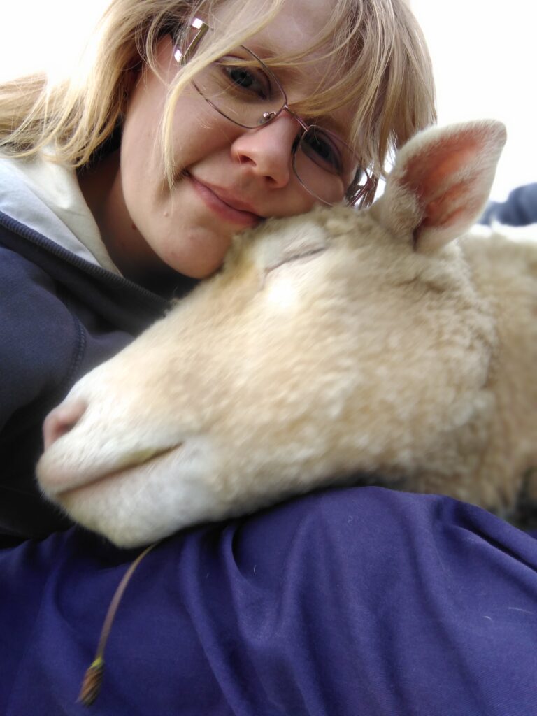 Blonde woman cuddling her pet sheep. The sheep is resting her head on the woman's shoulder and has her eyes closed. The sheep looks very content.