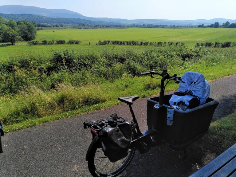 A cargo bike carrying a child in an infant car seat, on a sunny day. Glorious rolling countryside in the background.