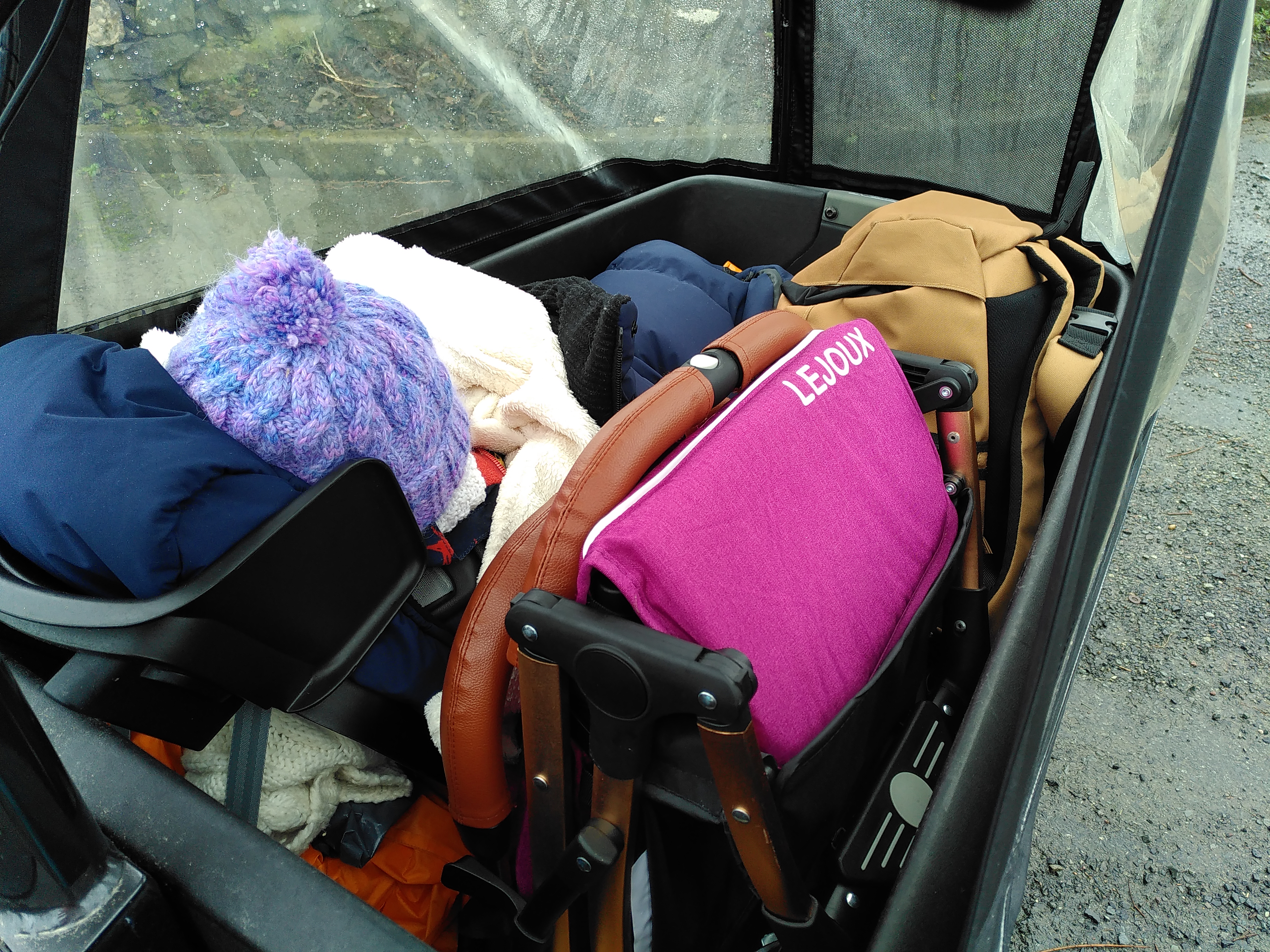 The bucket of a front-loader cargo bike. Within the bucket are a child in a child-seat, a compact buggy and changing bag.