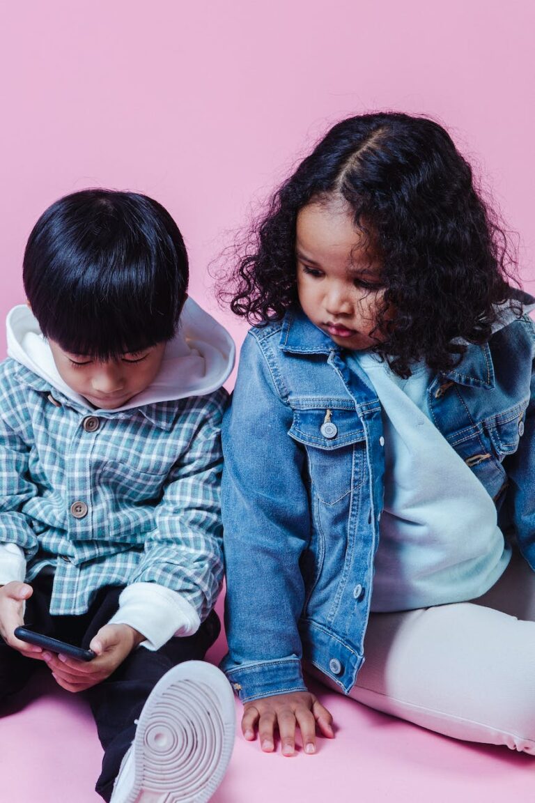 Two small children sit side-by-side. Both are fixated on a smart phone which the child on the right holds in his hands.