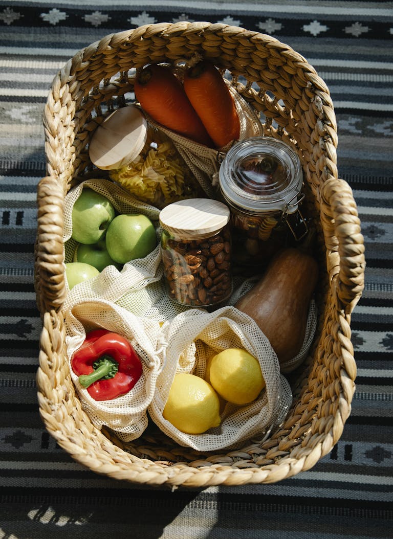 From above of wicker basket with vegetables and fruits placed in zero waste packaging with eco bags and glass jars with products on sunny day