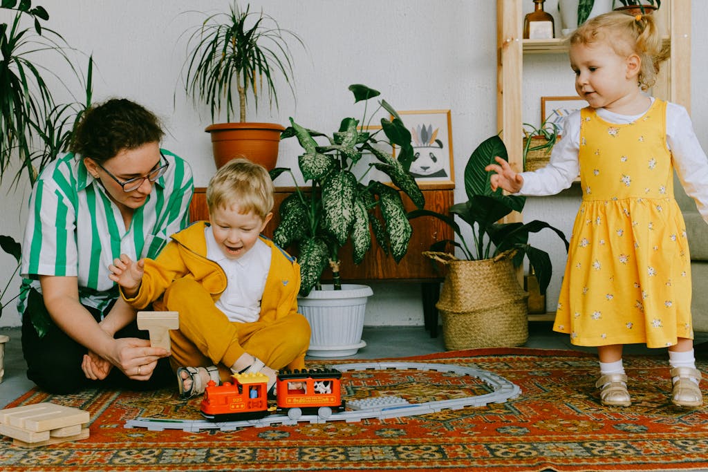 stay at home mum and adorable little brother and sister in casual wear gathering in cozy living room and having fun together while playing with plastic railway