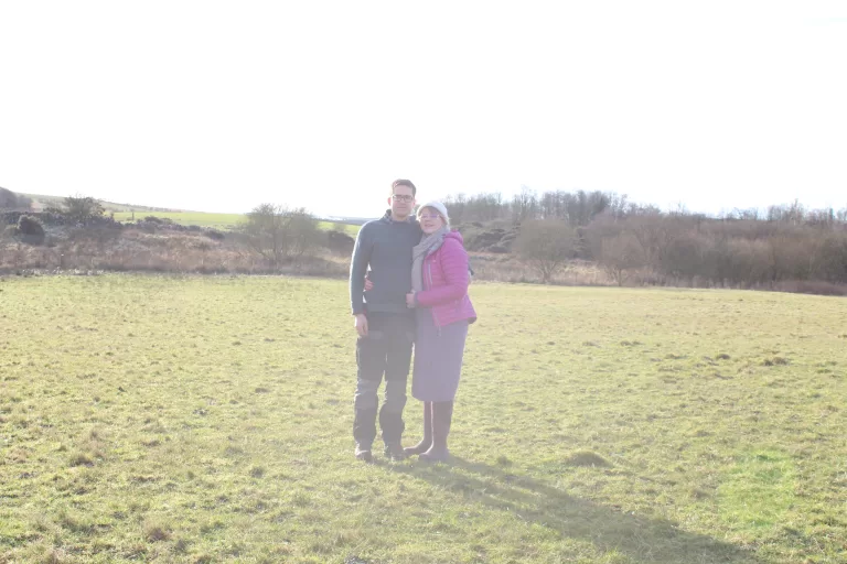 Man and woman dressed in warm clothing, standing in a green field on a sunny winter's day