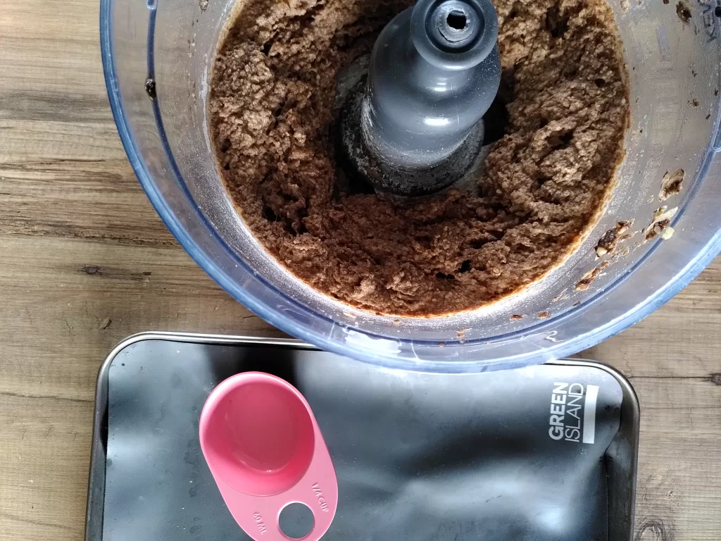 Food processor containing the mixture for nut burgers, next to a baking tray with a measuring cup and reusable baking liner.