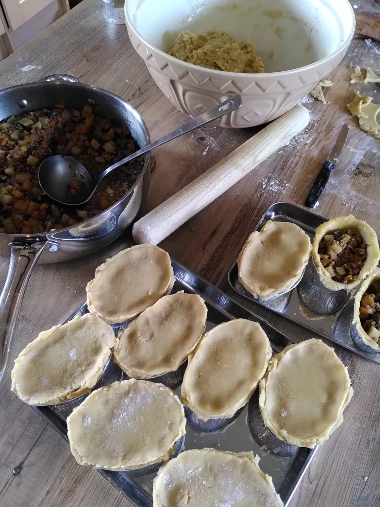 Batch-cooking home-made single-serve pies in a farmhouse kitchen. A large mixing bowl containing pastry, a rolling pin, and a saucepan with the pie filling.