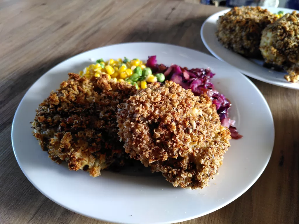 Fish-less cakes with peas, sweetcorn and red cabbage, served on a white china plate on a wooden counter top in a farmhouse kitchen.