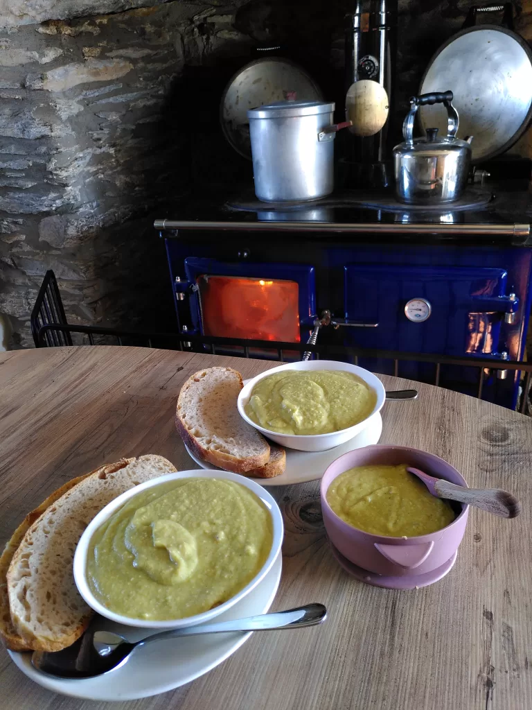 Three bowls of freshly made, creamy broccoli soup with fresh sourdough bread, situated in front of a wood burning range in a farmhouse kitchen.