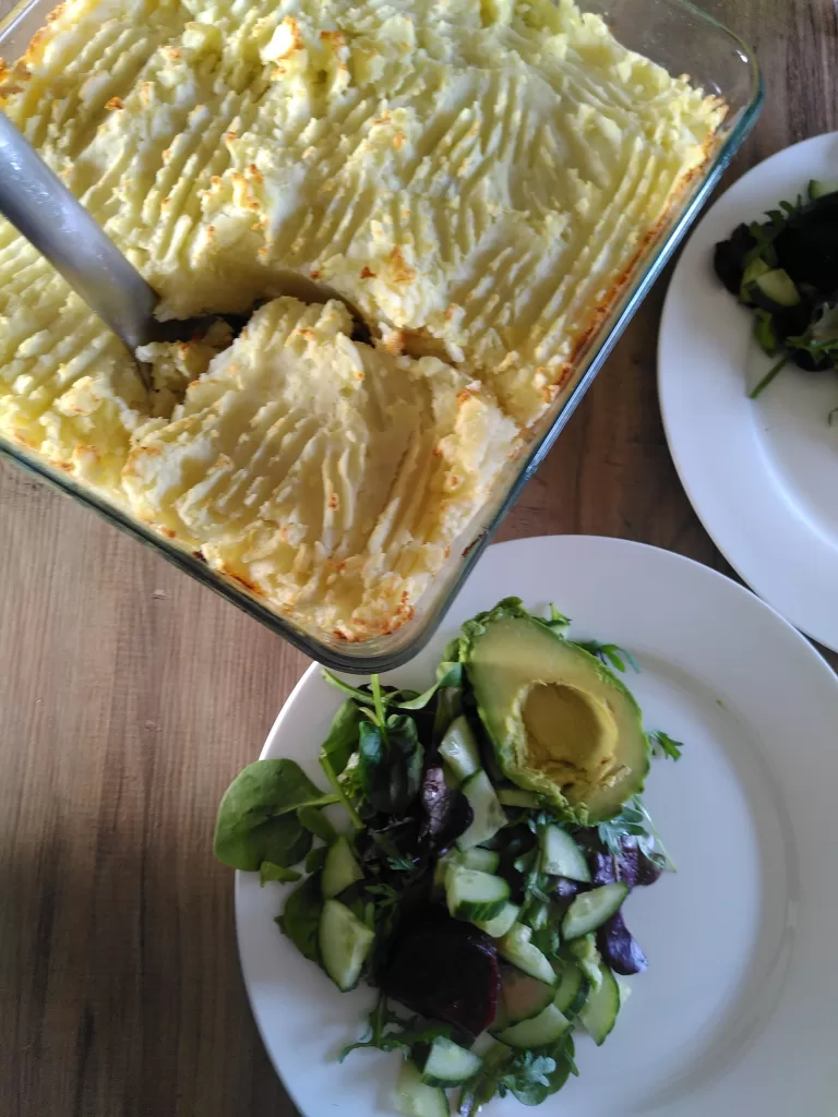 A dish containing vegan fishy pie, just out of the oven with a delectably crispy topping. The pie is ready to be dished up onto the plates beside it, both of which already have a green salad.