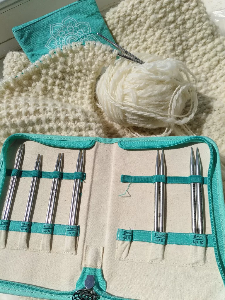 The open needle case of the KnitPro Believe interchangeable circular needles set, displayed alongside the accessory pouch and a knitting work in progress. The knitting project is of cream, home-spun yarn and is cast onto the 5mm needles of the aforementioned collection.